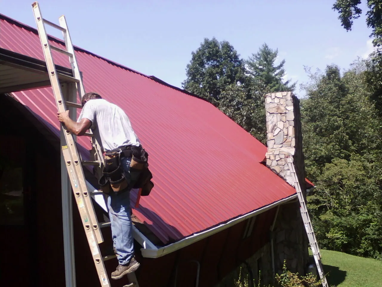 Roofer on a ladder beside a red metal roof and stone chimney — Soffit & Fascia Repair by Roofing Brothers in Weston
